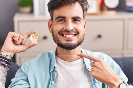 Foto de Joven hombre hispano con tatuajes sosteniendo la moneda criptomoneda sonriendo feliz señalando con la mano y el dedo - Imagen libre de derechos