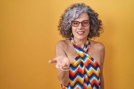 Foto de Mujer de mediana edad con el pelo gris de pie sobre fondo amarillo sonriente alegre que ofrece la palma de la mano dando asistencia y aceptación. - Imagen libre de derechos