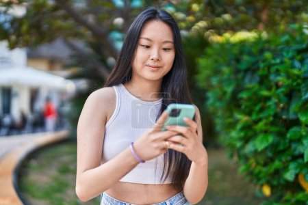 Téléchargez les photos : Jeune femme chinoise souriant confiant en utilisant smartphone au parc - en image libre de droit