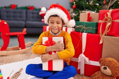 Foto de Adorable niño hispano abrazando regalo de Navidad sentado en el suelo en casa - Imagen libre de derechos