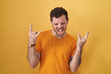 Photo for Young hispanic man standing over yellow background shouting with crazy expression doing rock symbol with hands up. music star. heavy music concept. - Royalty Free Image