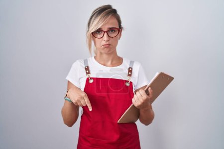 Photo for Young blonde woman wearing waiter uniform holding clipboard pointing down looking sad and upset, indicating direction with fingers, unhappy and depressed. - Royalty Free Image