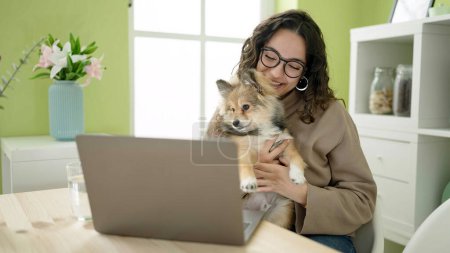 Photo for Young hispanic woman with dog sitting on table having video call at dinning room - Royalty Free Image