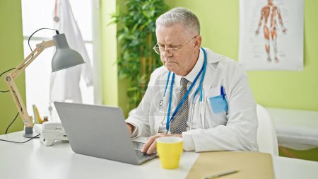 Téléchargez les photos : Moyen âge homme aux cheveux gris médecin utilisant un ordinateur portable à la clinique - en image libre de droit