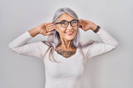 Foto de Mujer de mediana edad con el pelo gris de pie sobre fondo blanco sonriendo tirando de las orejas con los dedos, gesto divertido. problema de audición - Imagen libre de derechos