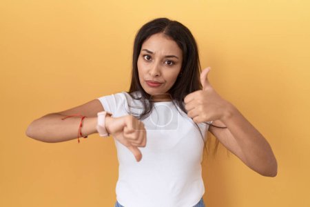 Photo for Young arab woman wearing casual white t shirt over yellow background doing thumbs up and down, disagreement and agreement expression. crazy conflict - Royalty Free Image