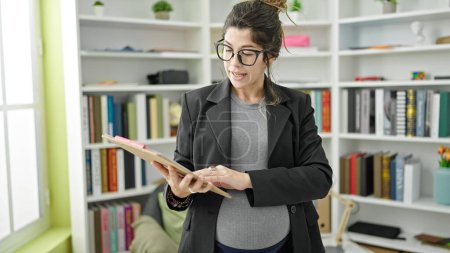 Téléchargez les photos : Jeune enseignante enceinte lisant le presse-papiers expliquant la leçon à la bibliothèque universitaire - en image libre de droit