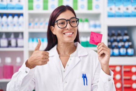 Téléchargez les photos : Jeune femme hispanique travaillant à la pharmacie tenant préservatif souriant heureux et positif, pouce levé faire excellent et signe d'approbation - en image libre de droit