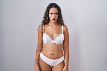 Téléchargez les photos : Jeune femme hispanique portant de la lingerie blanche dans le visage de choc, l'air sceptique et sarcastique, surpris avec la bouche ouverte - en image libre de droit