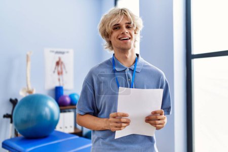 Foto de Joven pisioterapeuta hombre rubio sonriendo documento de lectura segura en la clínica de rehabilitación - Imagen libre de derechos