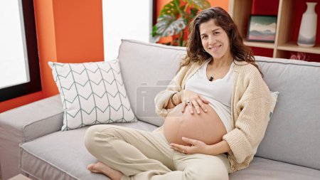 Téléchargez les photos : Jeune femme enceinte assise sur un canapé massant le ventre à la maison - en image libre de droit