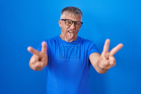 Téléchargez les photos : Homme hispanique avec les cheveux gris debout sur fond bleu souriant avec la langue montrant les doigts des deux mains faisant signe de victoire. numéro deux. - en image libre de droit