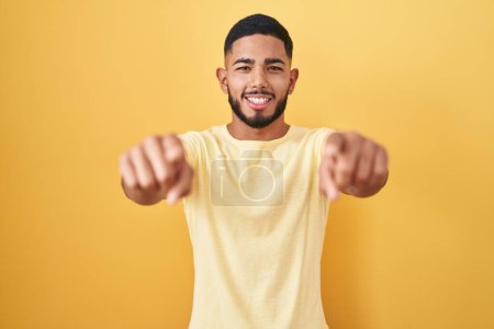 Foto de Young hispanic man standing over yellow background pointing to you and the camera with fingers, smiling positive and cheerful - Imagen libre de derechos