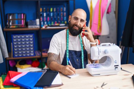 Téléchargez les photos : Jeune homme chauve tailleur parler sur smartphone écriture sur ordinateur portable à l'usine de vêtements - en image libre de droit