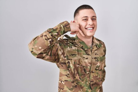 Foto de Joven vistiendo uniforme camuflaje del ejército sonriendo haciendo gesto telefónico con la mano y los dedos como hablando por teléfono. comunicar conceptos. - Imagen libre de derechos