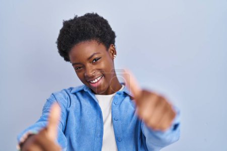 Photo for African american woman standing over blue background approving doing positive gesture with hand, thumbs up smiling and happy for success. winner gesture. - Royalty Free Image