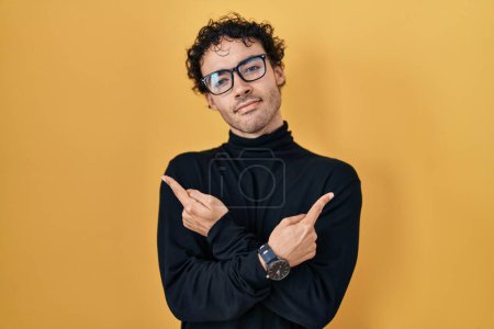 Photo for Hispanic man standing over yellow background pointing to both sides with fingers, different direction disagree - Royalty Free Image