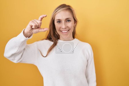 Photo for Young caucasian woman wearing white sweater over yellow background smiling and confident gesturing with hand doing small size sign with fingers looking and the camera. measure concept. - Royalty Free Image