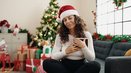 Téléchargez les photos : Moyen Age femme hispanique toucher le cœur avec photo assis sur le canapé par arbre de Noël à la maison - en image libre de droit