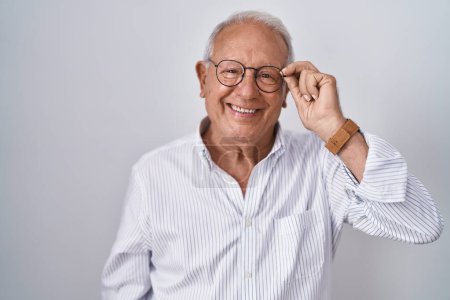 Foto de Hombre mayor con el pelo gris sosteniendo gafas con la mano mirando positiva y feliz de pie y sonriendo con una sonrisa confiada mostrando los dientes - Imagen libre de derechos