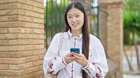 Photo for Young chinese woman using smartphone smiling at street - Royalty Free Image