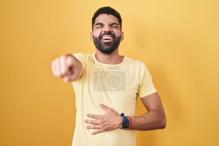 Téléchargez les photos : Homme hispanique avec la barbe debout sur fond jaune riant de vous, pointant du doigt la caméra avec la main sur le corps, expression de honte - en image libre de droit