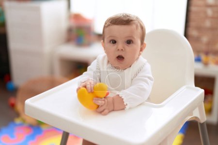 Photo for Adorable toddler sitting on baby highchair holding rubber duck toy at kindergarten - Royalty Free Image