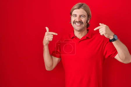 Foto de Hombre caucásico con bigote de pie sobre fondo rojo mirando confiado con sonrisa en la cara, señalándose con los dedos orgullosos y felices. - Imagen libre de derechos