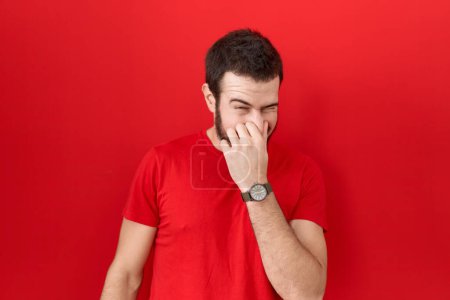 Foto de Joven hombre hispano usando una camiseta roja casual oliendo algo apestoso y asqueroso, olor intolerable, conteniendo la respiración con los dedos en la nariz. mal olor - Imagen libre de derechos