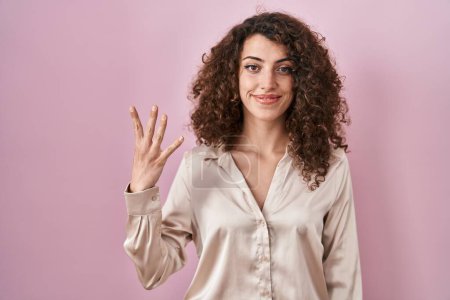 Foto de Mujer hispana con el pelo rizado de pie sobre fondo rosa mostrando y señalando hacia arriba con los dedos número cuatro mientras sonríe confiado y feliz. - Imagen libre de derechos