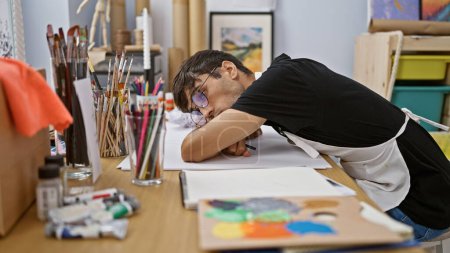 Foto de Joven hispano agotado, estudiante de arte, inmerso en su dibujo, se apoya en la mesa del estudio, luchando con la creatividad, rodeado de pinturas, pinceles y lienzos. - Imagen libre de derechos