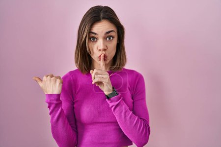 Téléchargez les photos : Femme hispanique debout sur fond rose demandant à être calme avec le doigt sur les lèvres pointant avec la main sur le côté. silence et concept secret. - en image libre de droit