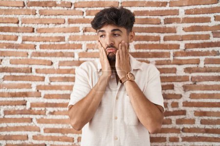 Photo for Arab man with beard standing over bricks wall background tired hands covering face, depression and sadness, upset and irritated for problem - Royalty Free Image