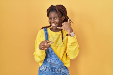 Téléchargez les photos : Femme africaine debout sur fond jaune souriant faire parler au téléphone geste et vous pointant du doigt. Appelle-moi. - en image libre de droit