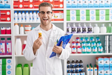 Photo for Young caucasian man working at pharmacy drugstore holding pills smiling and laughing hard out loud because funny crazy joke. - Royalty Free Image
