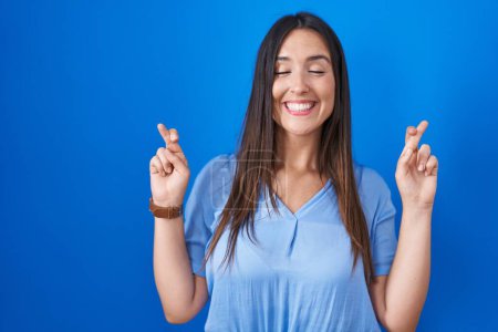 Téléchargez les photos : Jeune femme brune debout sur fond bleu geste doigt croisé souriant avec espoir et les yeux fermés. chance et concept superstitieux. - en image libre de droit