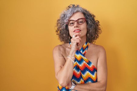 Photo for Middle age woman with grey hair standing over yellow background looking confident at the camera smiling with crossed arms and hand raised on chin. thinking positive. - Royalty Free Image