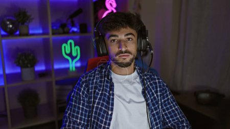 Photo for Focused in-game, young arab man, a serious-faced streamer, wearing headphones in a dim gaming room, engrossed in a futuristic virtual gaming world - Royalty Free Image