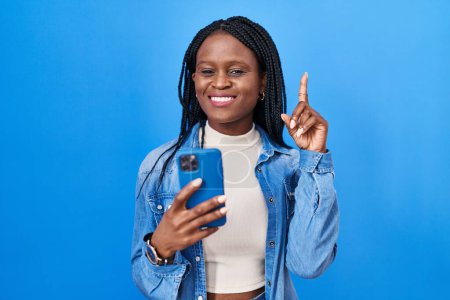 Téléchargez les photos : Femme africaine avec des tresses à l'aide d'un message de dactylographie smartphone surpris par une idée ou une question pointant du doigt avec un visage heureux, numéro un - en image libre de droit