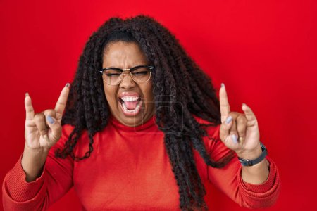 Photo for Plus size hispanic woman standing over red background shouting with crazy expression doing rock symbol with hands up. music star. heavy music concept. - Royalty Free Image