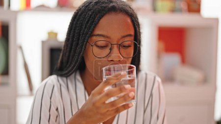 Téléchargez les photos : Femme afro-américaine buvant de l'eau assise sur la table à manger - en image libre de droit