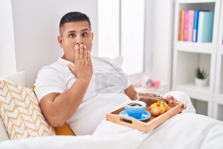 Foto de Joven hispano desayunando en la cama cubriendo la boca con la mano, sorprendido y temeroso de equivocarse. expresión sorprendida - Imagen libre de derechos
