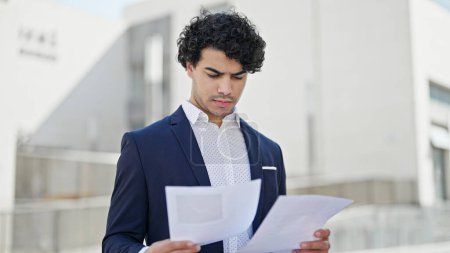 Téléchargez les photos : Jeune homme d'affaires latino-américain debout avec expression détendue document de lecture à la rue - en image libre de droit