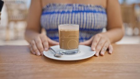 Photo for Young hispanic woman stirring coffee sitting on the table at sunny restaurant terrace - Royalty Free Image