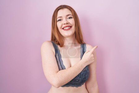 Photo for Redhead woman wearing lingerie over pink background cheerful with a smile on face pointing with hand and finger up to the side with happy and natural expression - Royalty Free Image