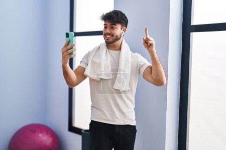 Foto de Hombre hispano con barba haciendo videollamadas con smartphone en el gimnasio sonriendo con una idea o pregunta apuntando con el dedo con la cara feliz, número uno - Imagen libre de derechos