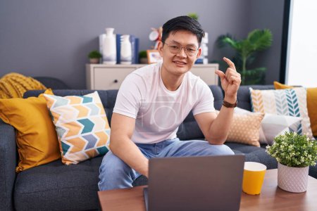 Foto de Joven hombre asiático usando el ordenador portátil en casa sentado en el sofá sonriente y seguro gesto con la mano haciendo signo de tamaño pequeño con los dedos mirando y la cámara. concepto de medida. - Imagen libre de derechos