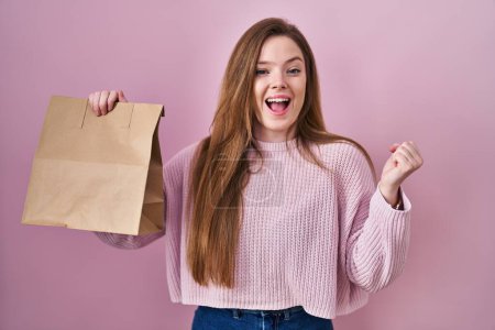 Photo for Young caucasian woman holding shopping bag and credit card screaming proud, celebrating victory and success very excited with raised arms - Royalty Free Image