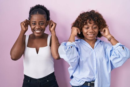 Téléchargez les photos : Deux femmes africaines debout sur fond rose souriant tirant les oreilles avec les doigts, geste drôle. problème d'audition - en image libre de droit