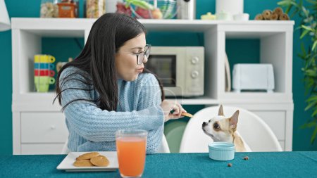 Photo for Young hispanic woman with chihuahua dog eating cookies sitting on the table at dinning room - Royalty Free Image